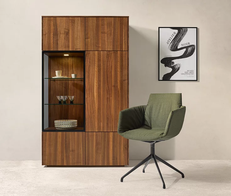 Elegant lui leger dining chair with olive-coloured Clara 793 fabric upholstery on a swivel base in matt black in front of a filigno display cabinet made of solid walnut and smoked glass
