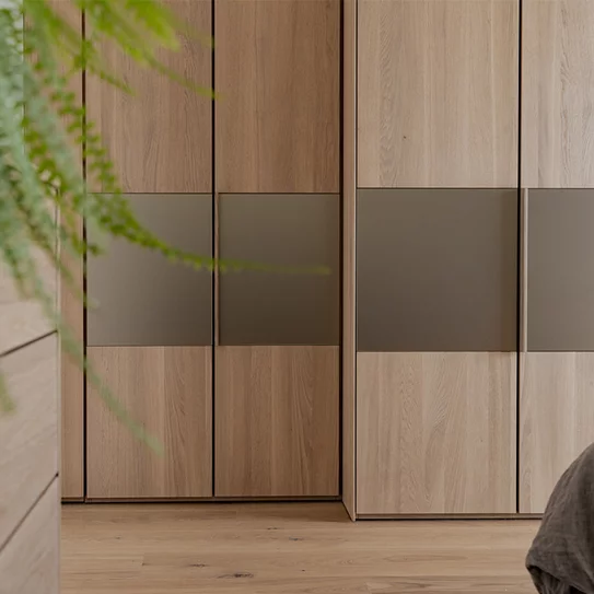 Massivholz Drehtuerenschrank valore in Eiche Weißöl mit integrierten Holzgriffleisten und Farbglaselementen in bronze