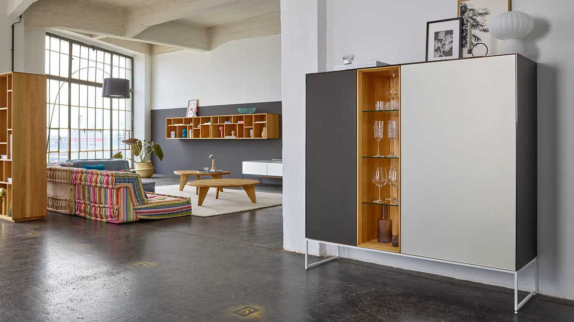 modern cubus pure wooden highboard made of solid oak with side panels, doors and top panel in coloured glass on a matt snow white metal frame, in the background two ur coffee tables and a graphic hanging shelf