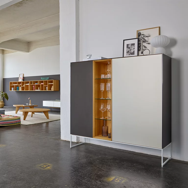 modern cubus pure wooden highboard made of solid oak with side panels, doors and top panel in coloured glass on a matt snow white metal frame, in the background two ur coffee tables and a graphic hanging shelf